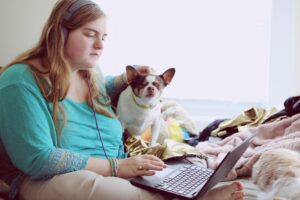 Young adult female wearing green top, sits on her bed staring at computer with her small dog sitting beside her. She looks sad.