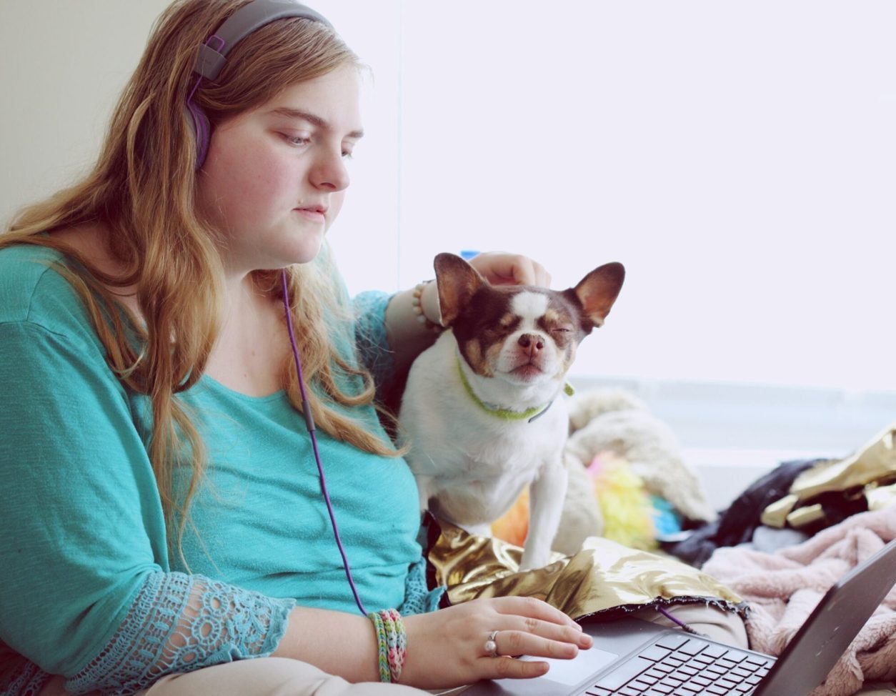 Young adult female wearing green top, sits on her bed staring at computer with her small dog sitting beside her. She looks sad.