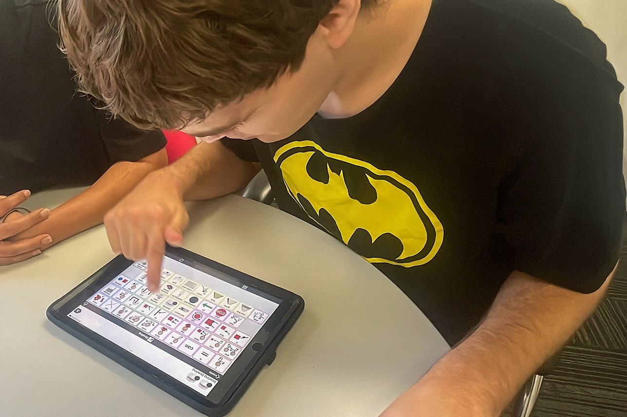 A male uses an Augmentative and Alternative Communication app on his ipad. He wears a black top and is intent on telling us about his day.