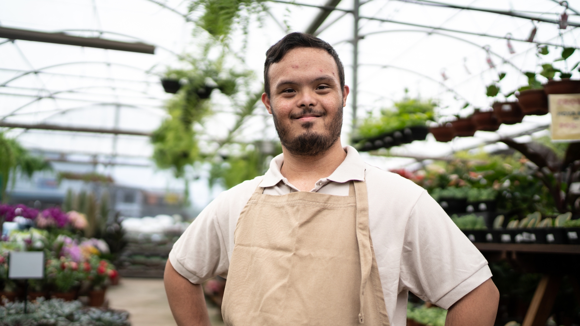 Young man standing inside a large commercial greehouse, smiling with hands on his hips. He is wearing a white polo top and a beige apron.