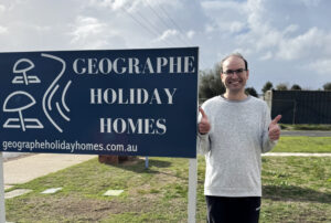 A person standing outdoors on a grassy area next to a large blue sign that reads “Geographe Holiday Homes” with the website address underneath. The person is giving two thumbs up, and the sky in the background is partly cloudy.